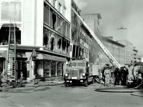 Schwarz-weiß-Foto eines Brands: Feuerwehr mit Leitern vor einem Haus mit geborstenen Fensterscheiben.