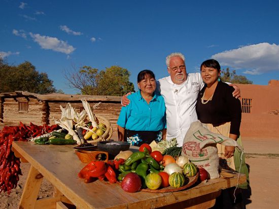 Ein Mann und zwei Frauen stehen Arm in Arm vor einem Tisch mit Gemüse und Obst.