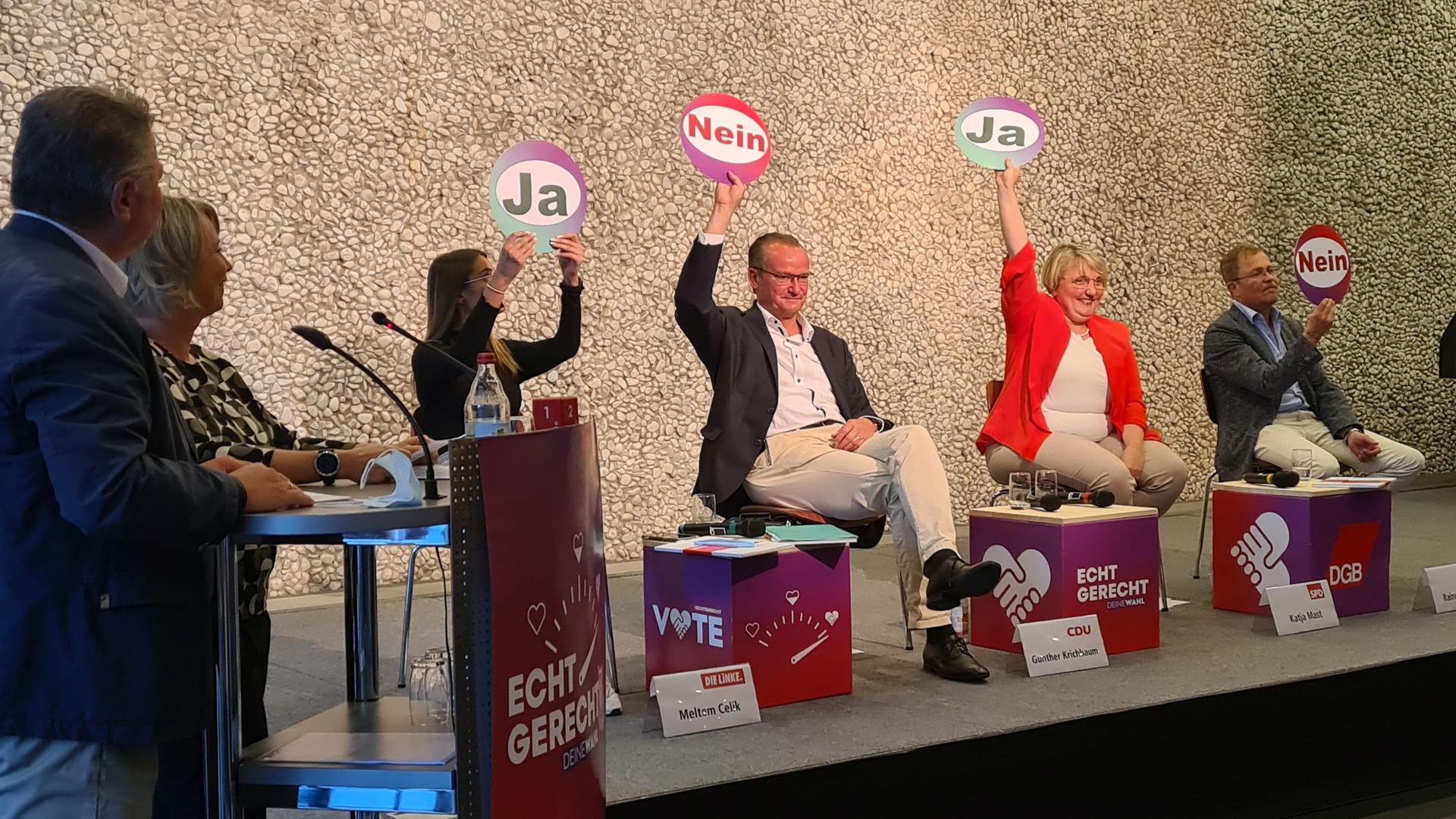 Links stehen zwei Personen an einem Podest, daneben sitzen die Bundestagskandidatinnen und -kandidaten, sie halten Schilder hoch, auf denen „Ja“ oder „Nein“ steht