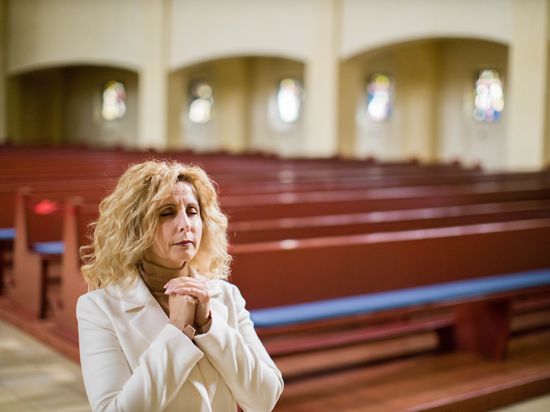 Eine Frau steht mit gefalteten Händen im Innenraum einer Kirche. Im Hintergrund sind Kirchenbänke.