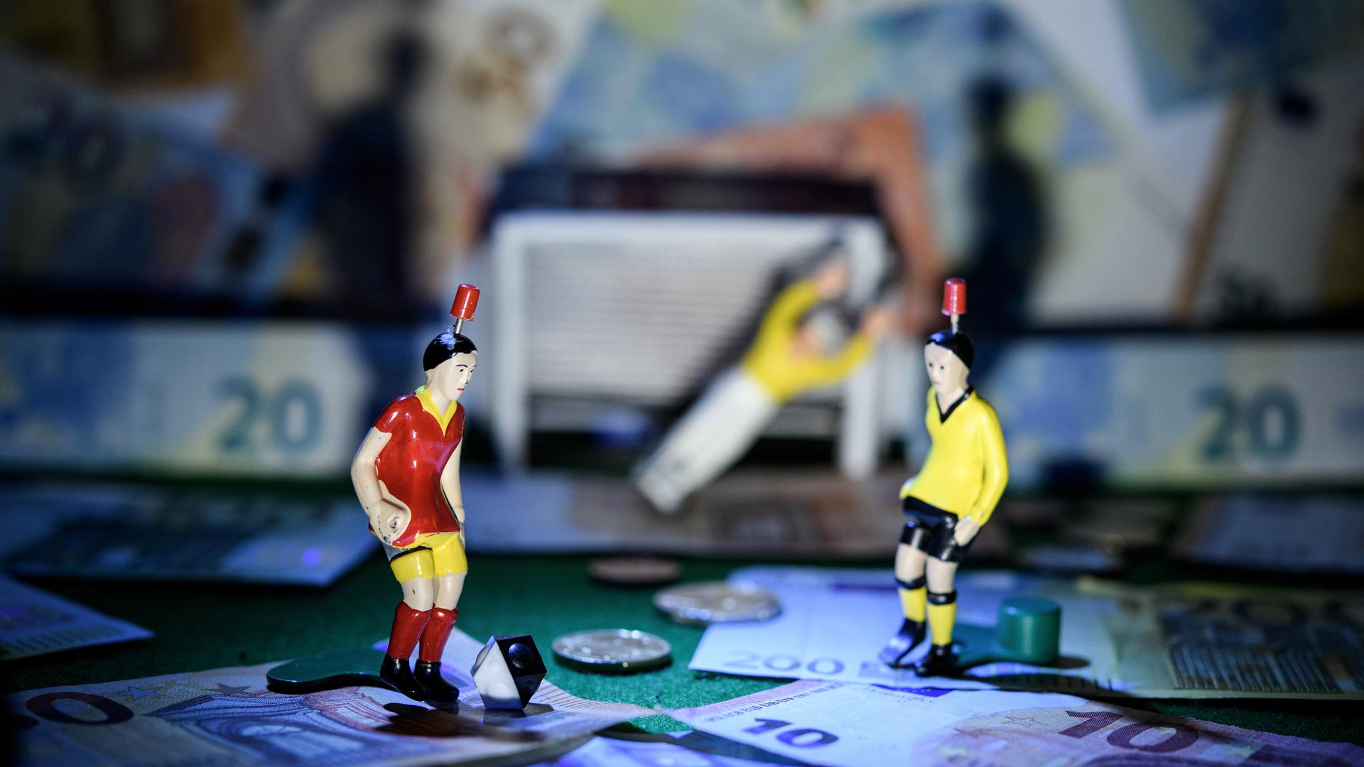 Männchen stehen auf einem Tipp-Kick-Spielfeld, auf und neben dem zahlreiche Geldscheine zu sehen sind.
