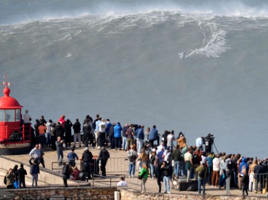 Menschen am Leuchtturm bei Nazaré beobachten einen Surfer am Praia do Norte, der eine Riesenwelle surft.
